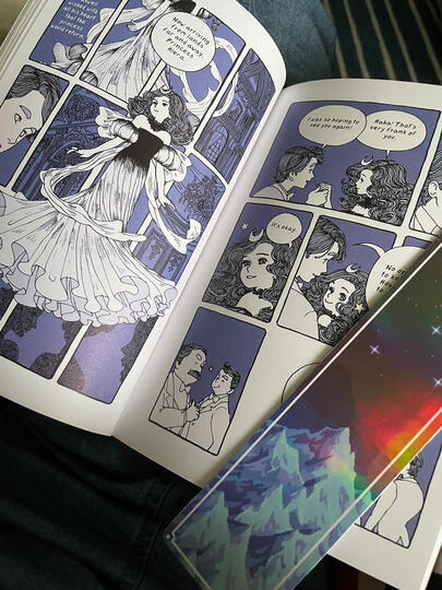The Magic Fish The Magic Fish graphic novel by Trung opened up showing several pages with beautiful illustrations of characters in fancy outfits, an iridescent bookmark of a mountain is laying across one page.