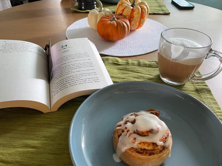 Legends & Lattes Legends & Lattes book open on a table next to coffee and a cinnamon roll on a plate.
