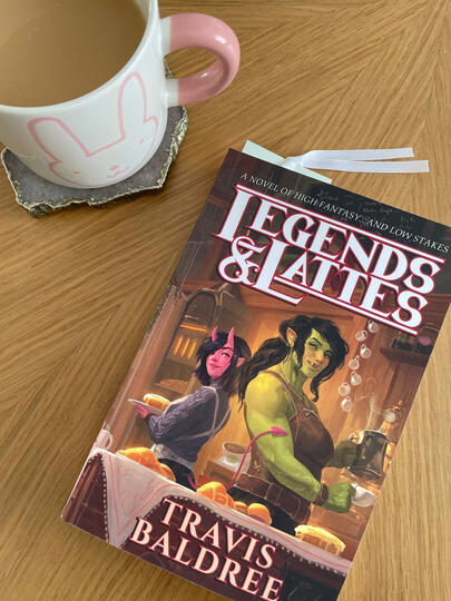 Legends & Lattes Legends & Lattes book by Travis Baldree sitting on a table next to a latte in a mug.
